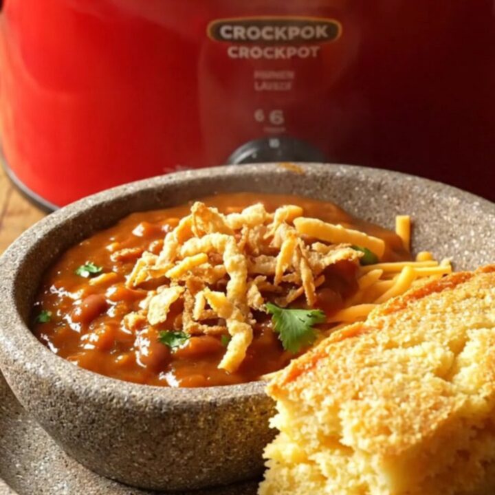 A close-up shot of a deep red ceramic slow cooker filled with glossy, sweet-heat BBQ chicken chili with melted cheese.