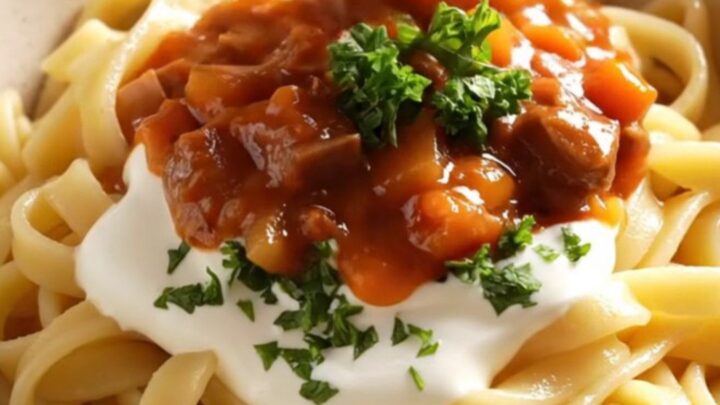 A close-up of a copper-red ceramic crockpot filled with rich, crimson beef goulash and fresh parsley.