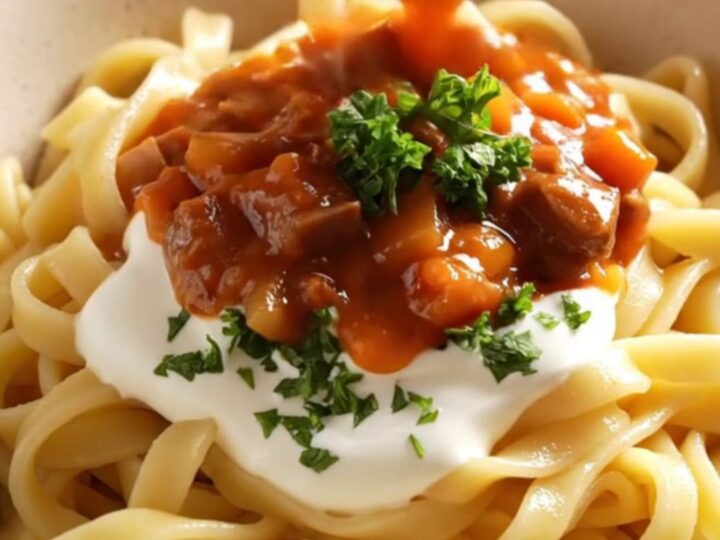 A close-up of a copper-red ceramic crockpot filled with rich, crimson beef goulash and fresh parsley.
