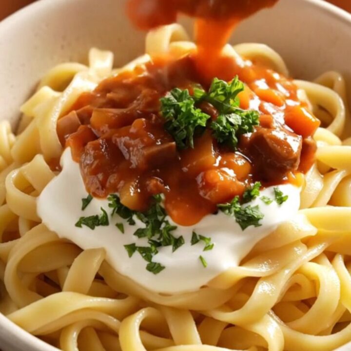 A close-up of a copper-red ceramic crockpot filled with rich, crimson beef goulash and fresh parsley.