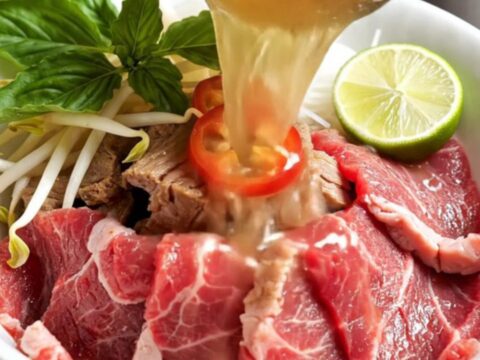 A steaming bowl of Vietnamese beef pho with brisket, noodles, and fresh herbs in a dark ceramic bowl.