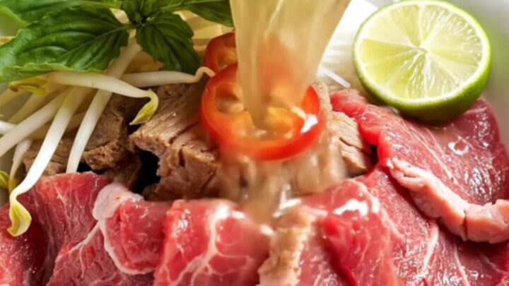 A steaming bowl of Vietnamese beef pho with brisket, noodles, and fresh herbs in a dark ceramic bowl.