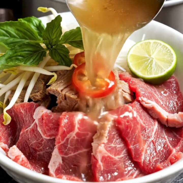 A steaming bowl of Vietnamese beef pho with brisket, noodles, and fresh herbs in a dark ceramic bowl.