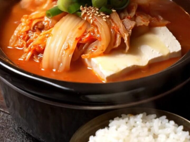 A steaming bowl of vibrant red kimchi jjigae topped with fresh scallions and sesame seeds.