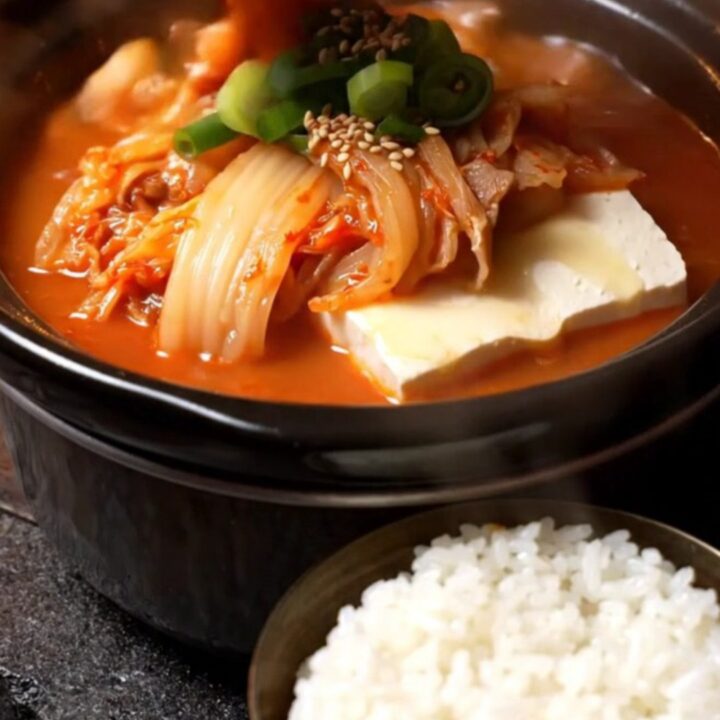 A steaming bowl of vibrant red kimchi jjigae topped with fresh scallions and sesame seeds.