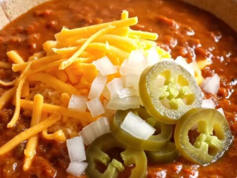 A warm stoneware bowl filled with deep red Texas chili con carne, topped with sharp cheddar, onion, and jalapenos.