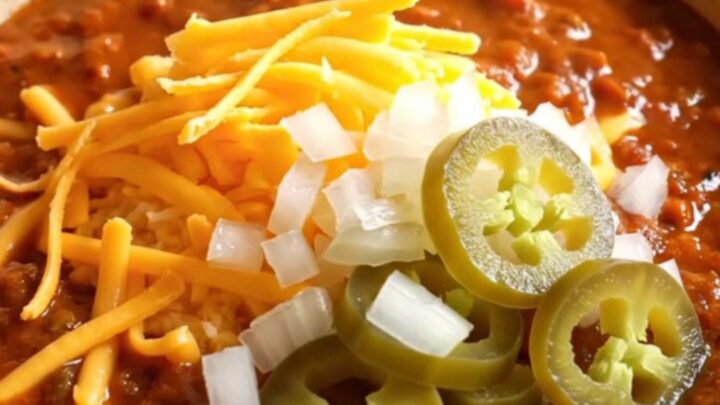 A warm stoneware bowl filled with deep red Texas chili con carne, topped with sharp cheddar, onion, and jalapenos.
