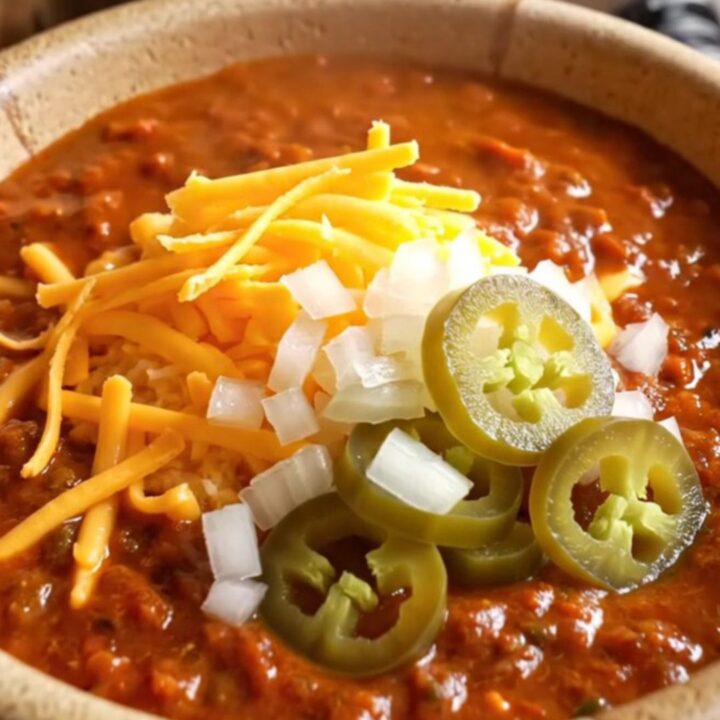A warm stoneware bowl filled with deep red Texas chili con carne, topped with sharp cheddar, onion, and jalapenos.