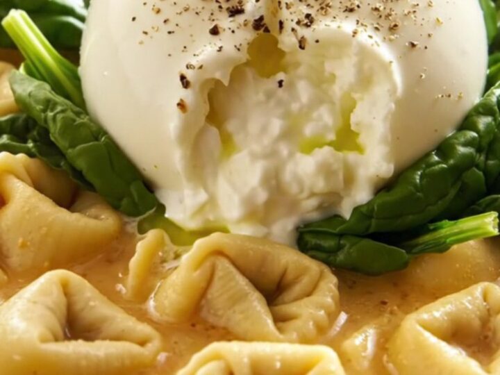 A close-up of a bowl of creamy tortellini soup topped with a whole, fresh, creamy burrata ball and green basil oil.