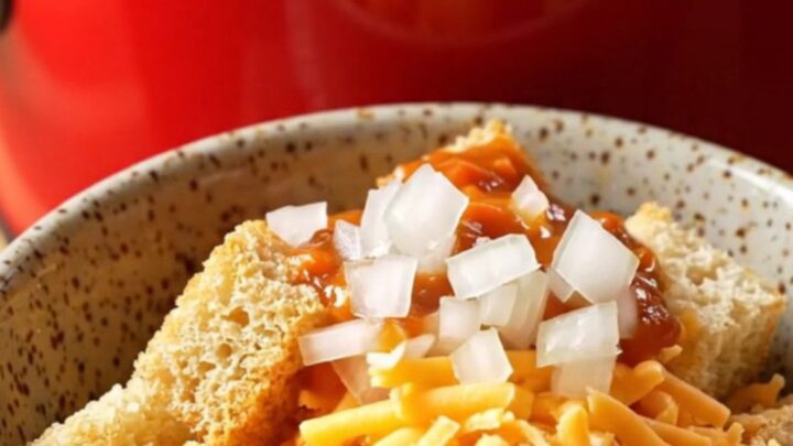 A ceramic slow cooker filled with creamy chili cheese dog soup topped with shredded cheddar and diced onions.