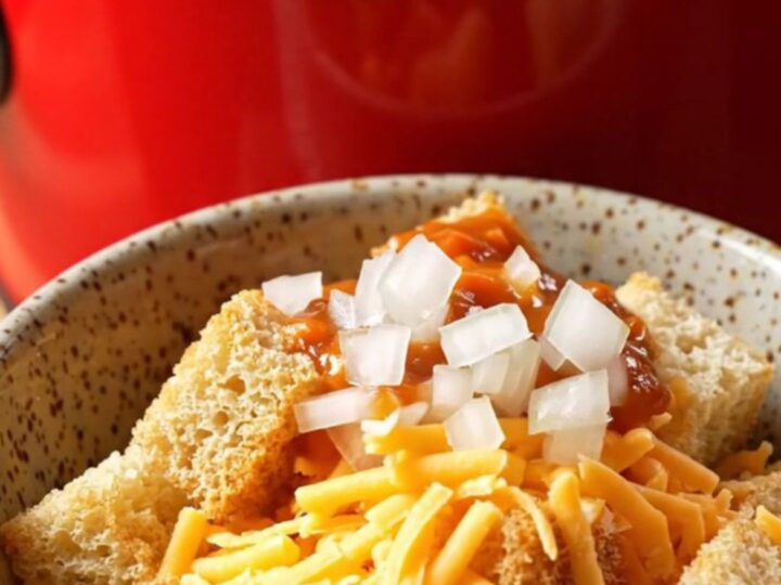 A ceramic slow cooker filled with creamy chili cheese dog soup topped with shredded cheddar and diced onions.