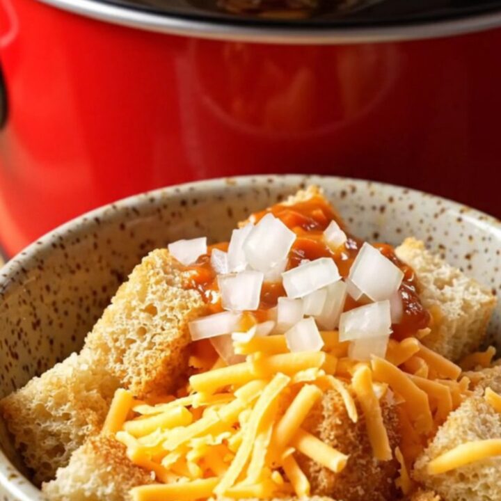 A ceramic slow cooker filled with creamy chili cheese dog soup topped with shredded cheddar and diced onions.