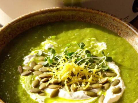 A warm ceramic bowl filled with vibrant green vegetable soup, topped with a mascarpone swirl, toasted pepitas