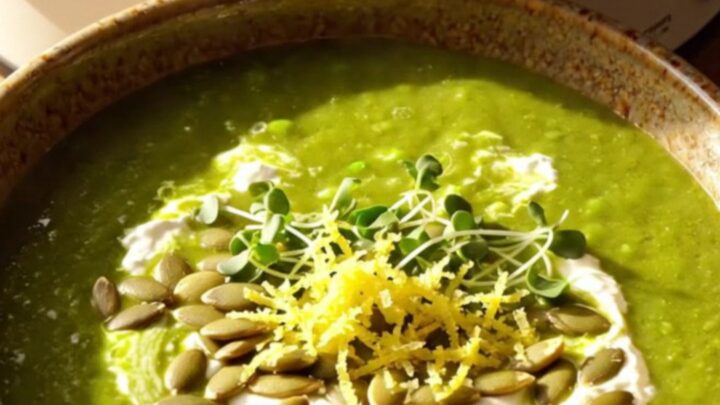 A warm ceramic bowl filled with vibrant green vegetable soup, topped with a mascarpone swirl, toasted pepitas