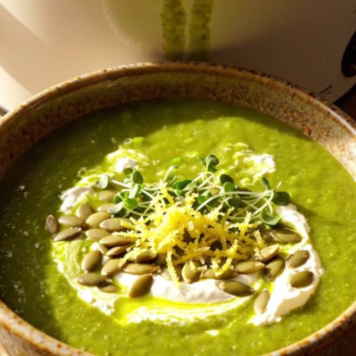 A warm ceramic bowl filled with vibrant green vegetable soup, topped with a mascarpone swirl, toasted pepitas