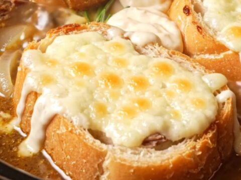 A deep stoneware crock filled with rich French dip soup topped with a golden, bubbly Gruyere cheese baguette slice.