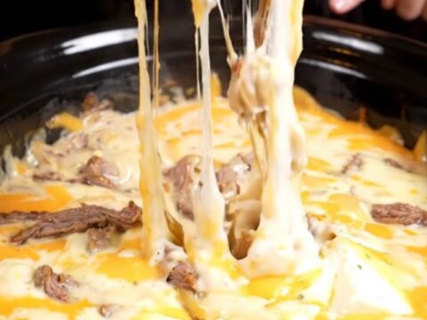 A close-up of a toasted baguette slice dipping into a bubbling, creamy Philly cheesesteak queso dip with visible ribeye.
