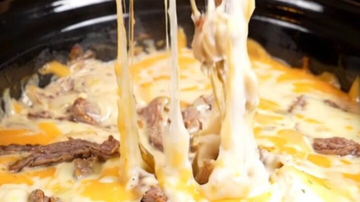 A close-up of a toasted baguette slice dipping into a bubbling, creamy Philly cheesesteak queso dip with visible ribeye.