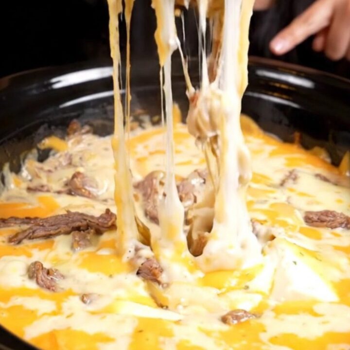 A close-up of a toasted baguette slice dipping into a bubbling, creamy Philly cheesesteak queso dip with visible ribeye.