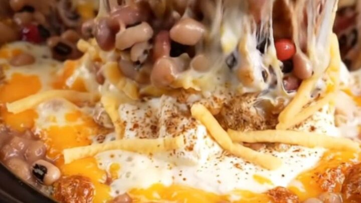 A macro shot of a tortilla chip scooping into a bubbling, cheesy, bean-filled dip inside a red slow cooker.