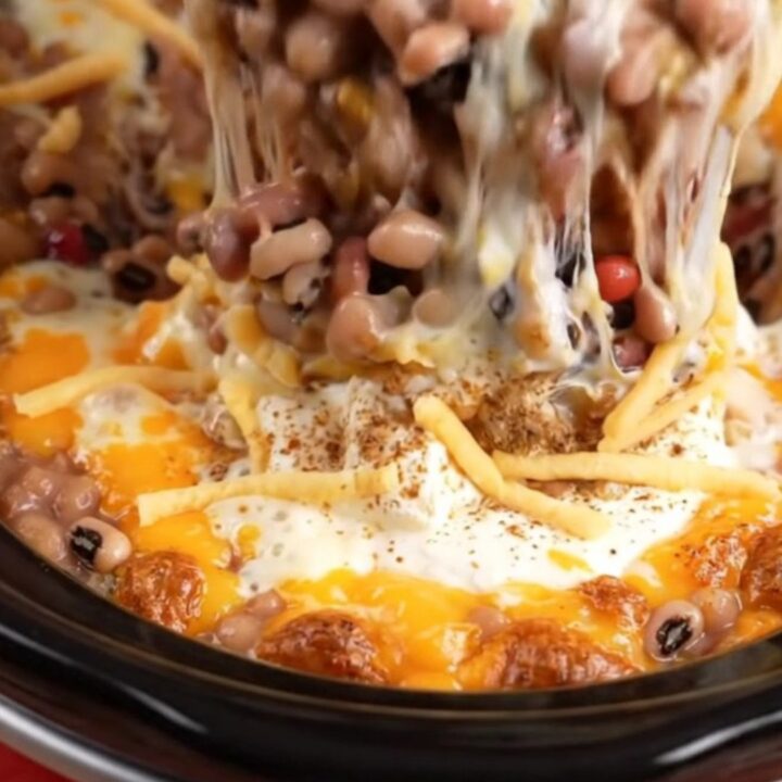 A macro shot of a tortilla chip scooping into a bubbling, cheesy, bean-filled dip inside a red slow cooker.