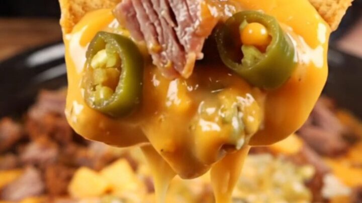 A close-up shot of a thick tortilla chip being dipped into a creamy, brisket-loaded queso dip inside a black slow cooker.