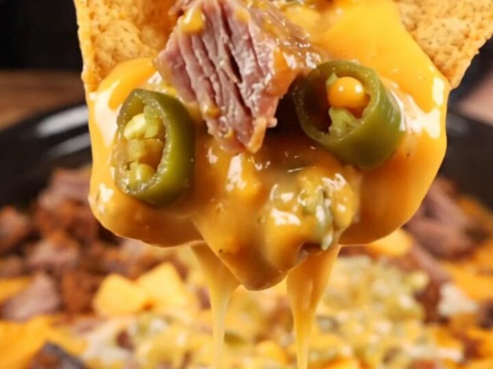 A close-up shot of a thick tortilla chip being dipped into a creamy, brisket-loaded queso dip inside a black slow cooker.