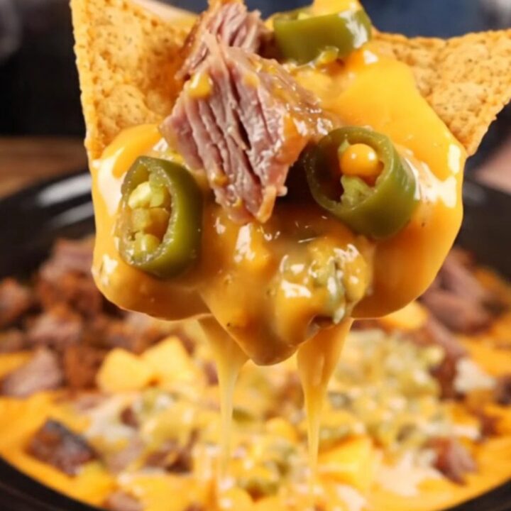 A close-up shot of a thick tortilla chip being dipped into a creamy, brisket-loaded queso dip inside a black slow cooker.