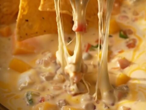 A close-up of a tortilla chip being dipped into a creamy, bubbling mango habanero queso in a white ceramic crockpot.