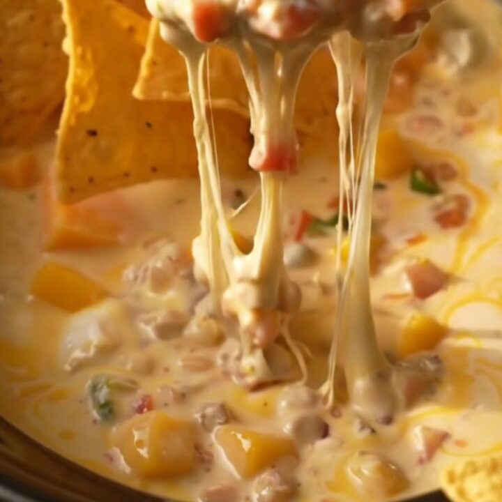 A close-up of a tortilla chip being dipped into a creamy, bubbling mango habanero queso in a white ceramic crockpot.