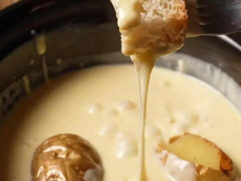 A ceramic slow cooker filled with creamy, melted Swiss and Gruyere fondue, with a rustic bread cube being dipped into it.