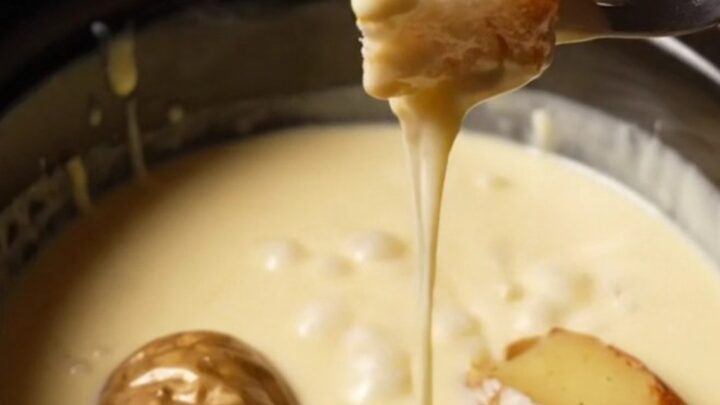 A ceramic slow cooker filled with creamy, melted Swiss and Gruyere fondue, with a rustic bread cube being dipped into it.