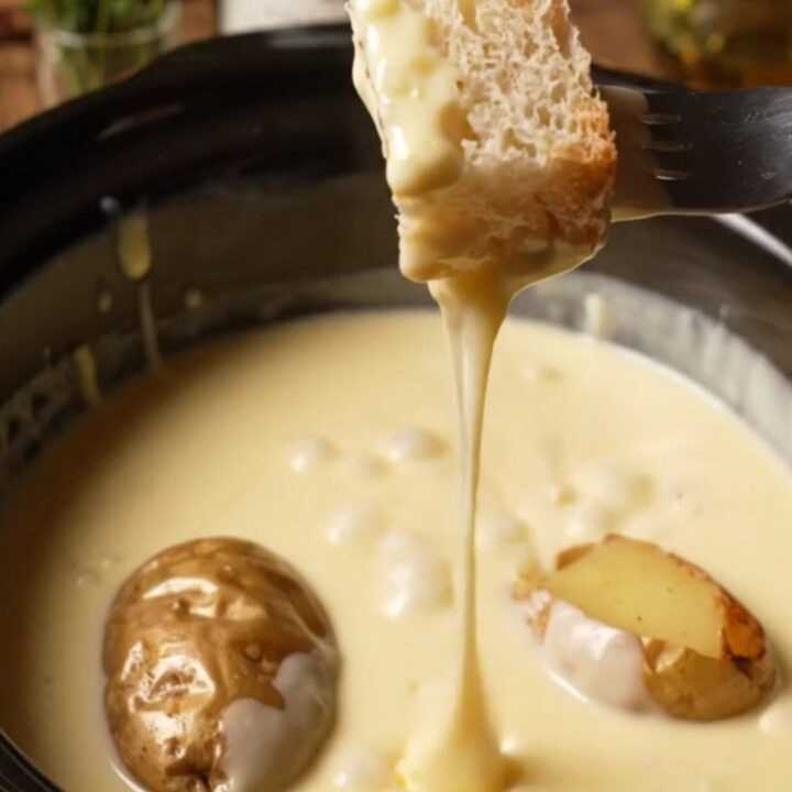 A ceramic slow cooker filled with creamy, melted Swiss and Gruyere fondue, with a rustic bread cube being dipped into it.