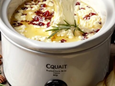 A close-up of a cracker dipping into a bubbling crockpot of melted brie with cranberries and pecans.