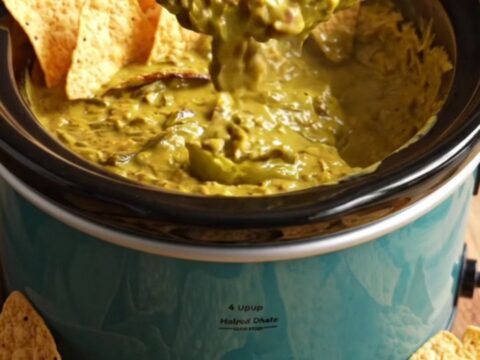A close-up of a corn tortilla chip being dipped into a bubbling, golden-green Hatch chile and smoked cheddar queso dip.