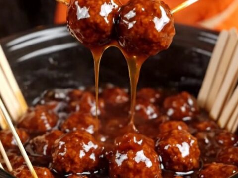 A close-up of a meatball glazed in sticky sauce being lifted from a black slow cooker with a bamboo pick.