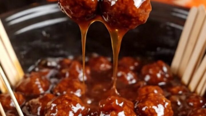 A close-up of a meatball glazed in sticky sauce being lifted from a black slow cooker with a bamboo pick.