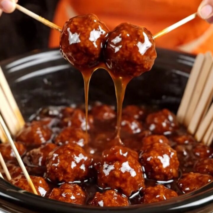 A close-up of a meatball glazed in sticky sauce being lifted from a black slow cooker with a bamboo pick.