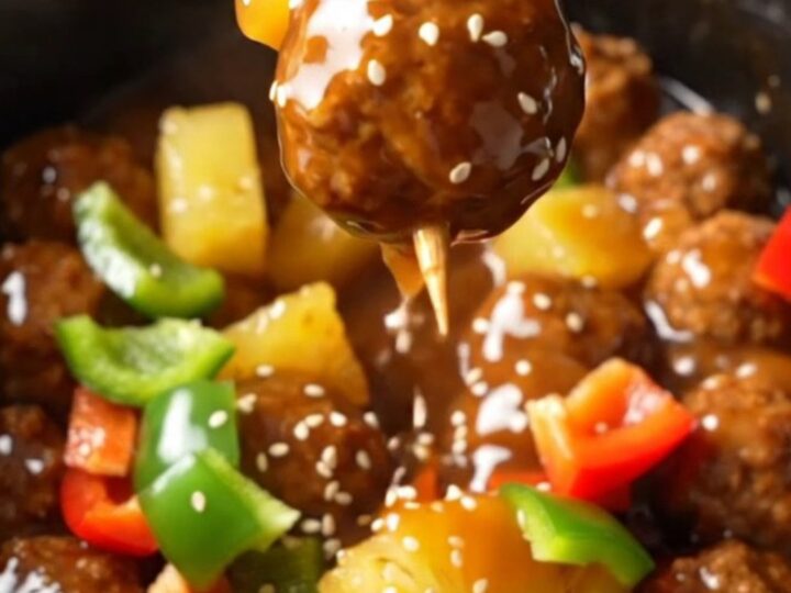 A close-up of glossy sweet and sour Hawaiian meatballs with pineapple and peppers in a black slow cooker.