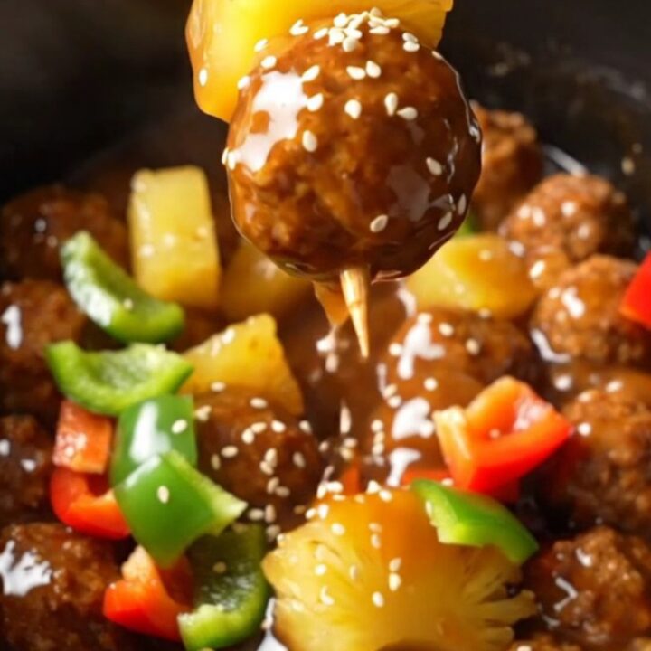 A close-up of glossy sweet and sour Hawaiian meatballs with pineapple and peppers in a black slow cooker.