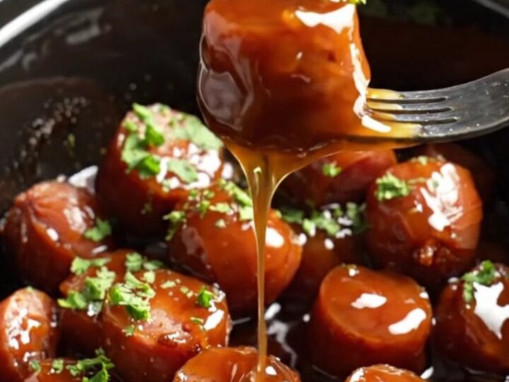 Close-up of sticky bourbon maple glazed kielbasa bites in a dark ceramic slow cooker, garnished with green parsley.