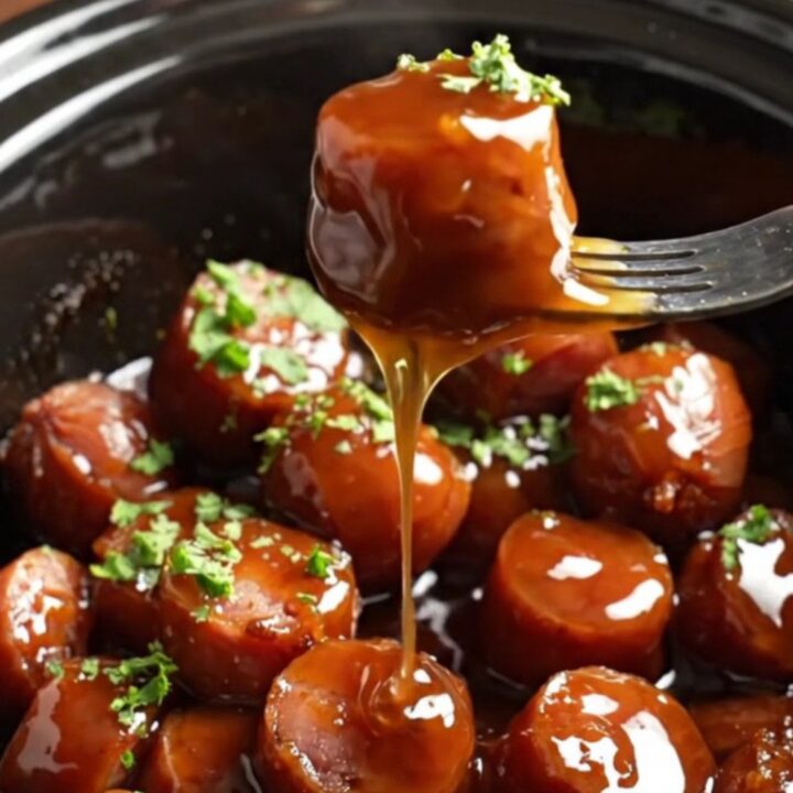Close-up of sticky bourbon maple glazed kielbasa bites in a dark ceramic slow cooker, garnished with green parsley.