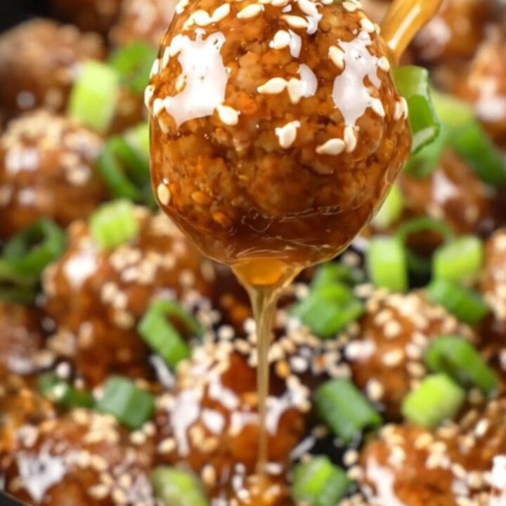 A close-up of sticky honey garlic meatballs in a black ceramic slow cooker, garnished with sesame seeds and scallions.