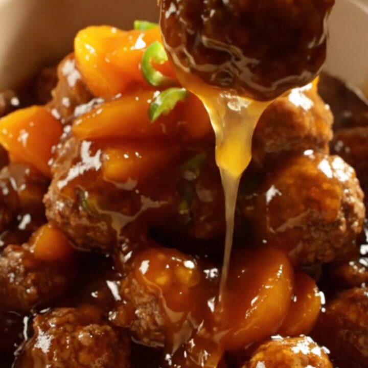 A close-up of golden-amber glazed meatballs in a cream stoneware crockpot, with fruit-studded BBQ sauce dripping down.
