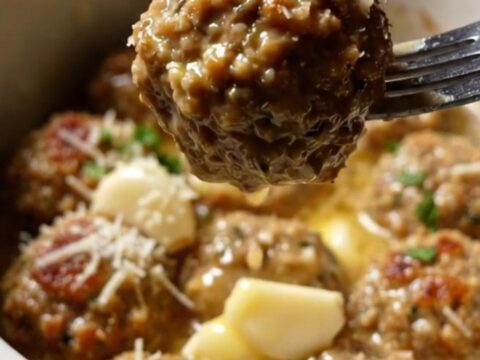 A golden herb-crusted chicken meatball being lifted from a white ceramic Crockpot filled with garlic butter sauce.