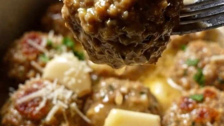 A golden herb-crusted chicken meatball being lifted from a white ceramic Crockpot filled with garlic butter sauce.