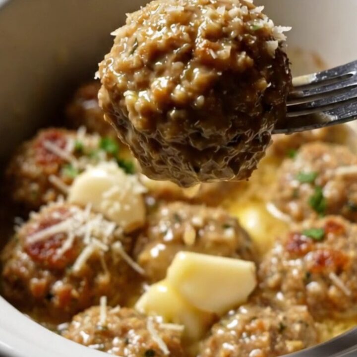 A golden herb-crusted chicken meatball being lifted from a white ceramic Crockpot filled with garlic butter sauce.