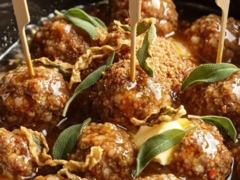 A ceramic slow cooker filled with amber-glazed pork meatballs garnished with fresh sage leaves.