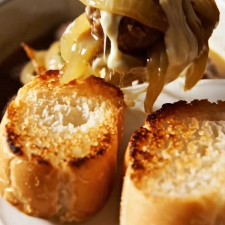 A close-up of tender beef meatballs in a ceramic slow cooker, topped with melted, gooey Gruyere cheese and fresh thyme.