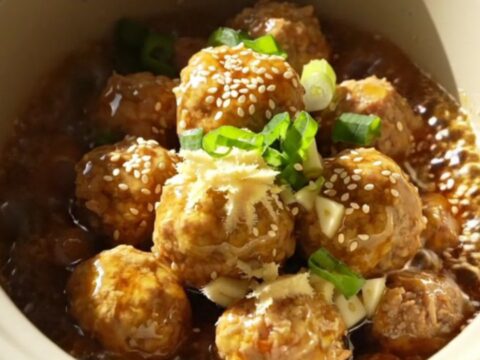 A close-up of golden-amber glazed meatballs in a ceramic slow cooker, garnished with green scallions.